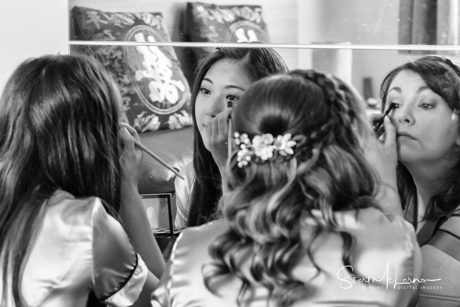 The bride and her bridesmaid share a bedroom mirror as they apply makeup