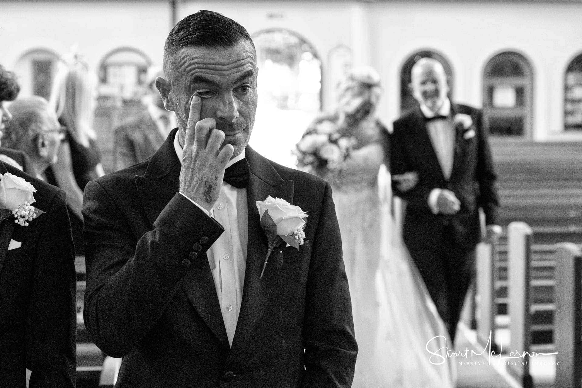 The groom wipes a tear from his eye as his bride walks up the aisle